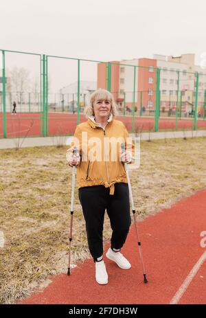Une femme âgée dans une veste jaune se tient avec des bâtons nordiques dans les mains au stade sur un tapis roulant rouge. Mode de vie sain Banque D'Images