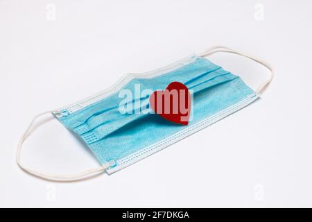 Un masque médical bleu avec un petit coeur en feutre rouge sur ses plis, 2021 février, COVID-19 thème de la Saint-Valentin. Banque D'Images