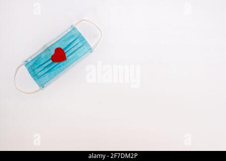 Un masque médical bleu avec un petit coeur en feutre rouge sur ses plis, 2021 février, COVID-19 thème de la Saint-Valentin (salle pour texte, copie). Banque D'Images