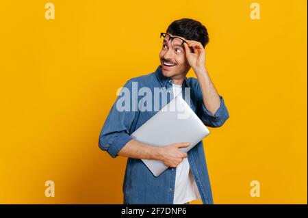 Un homme charismatique émerveillé avec des lunettes et élégamment habillé, tient un ordinateur portable à portée de main, semble surpris par l'espace vide prenant des lunettes, sourire, isolé arrière-plan orange, espace de copie Banque D'Images
