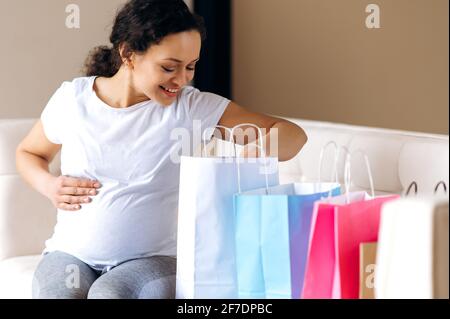 Femme enceinte heureuse, femme de course mixte, assise à la maison sur le canapé, dans des vêtements décontractés, regardant dans des sacs en papier avec de nouveaux vêtements ou jouets pour le futur bébé, souriant, concept de shopping Banque D'Images