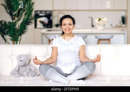 Belle heureuse adorable future maman, brunette, course mixte, assis en position lotus sur le canapé à la maison dans le salon, détente et méditer, regardant l'appareil photo, souriant mignon Banque D'Images