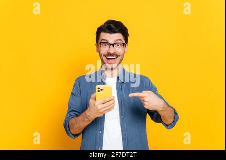 Joyeux caucasien stylisé gars avec des lunettes, en tenant le smartphone à la main et en le pointant vers le doigt, regardant avec plaisir l'appareil photo avec sourire, debout sur un arrière-plan isolé orange Banque D'Images