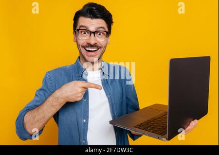 Joyeux et joyeux caucasien stylisé gars avec des lunettes, tient un ordinateur portable ouvert dans sa main, le pointant vers elle avec le doigt, regarde stupéfié à la caméra, se tient sur un arrière-plan isolé orange Banque D'Images