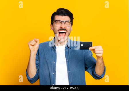 Joyeux caucasien gai unshaven stylisé gars tenant la carte de crédit à la main, criant joyeusement, debout sur un fond orange isolé, finance et le concept bancaire Banque D'Images