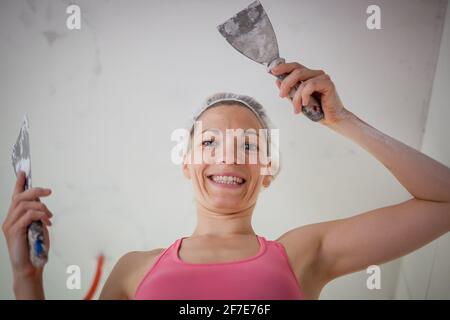 Jeune femme portant un dessus rose et tenant deux spatules à la main tout en travaillant avec le plâtre pendant la rénovation d'un appartement. Bonne femme de race blanche reno Banque D'Images