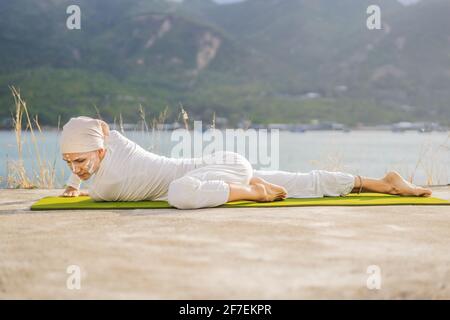 Kundalini yoga femme en vêtements blancs et turban pratique le yoga kundalini sur le fond de la mer, des montagnes et du coucher du soleil. Peinture de visage de combat Banque D'Images