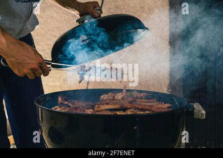 Homme ouvrant un barbecue fumeur ou un four à charbon de bois, pour faire tourner des kebabs ou des côtes de rechange au gril sur la chaleur. Fumée visible s'élevant du gril Banque D'Images