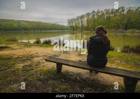 Femme assise sur le banc sur les rives de Bukovnisko jezero ou au lac de Prekmurje, en Slovénie, un jour de printemps froid et couvert. Banque D'Images