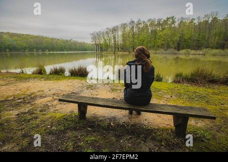 Femme assise sur le banc sur les rives de Bukovnisko jezero ou au lac de Prekmurje, en Slovénie, un jour de printemps froid et couvert. Banque D'Images