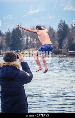 Un inconnu saute dans un lac froid de Bled, en Slovénie en hiver, une personne en premier plan est en train de filmer ou de prendre des photos. Banque D'Images