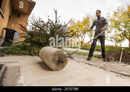 Un homme utilise un vieux rouleau en béton pour durcir le sol recouvert de sable devant une maison résidentielle. Banque D'Images