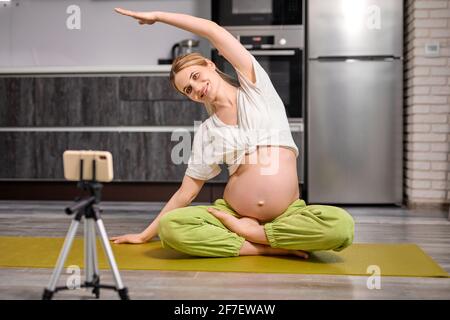 Femme enceinte agréable faire des exercices de yoga regarder la vidéo en ligne sur smartphone , s'asseoir sur le sol Performing Exercises, train flexibilité, à la maison. Banque D'Images