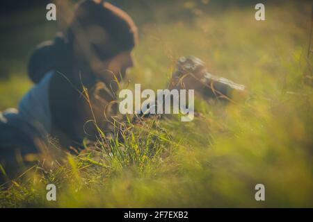 Un homme avec une casquette et photocamera, un photographe se cachant dans l'herbe. Concentrez-vous sur une masse d'herbe avec un photographe qui vérifie les photos en arrière-plan Banque D'Images