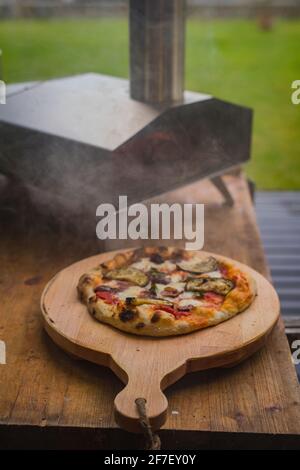 Délicieuse pizza maison fraîche sur une assiette ronde en bois, directement à partir d'un four à bâble maison en acier inoxydable alimenté par des granulés de bois. Pizza en plein air. Banque D'Images