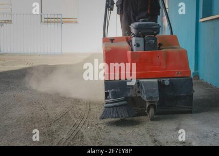 Une personne pousse un balai industriel à essence orange sur un sol en asphalte sale pour tenter de le nettoyer des débris et du sable Banque D'Images