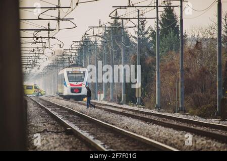 Homme qui marche de manière ignorante sur les voies de train avec train de passagers blanc à l'approche. Une ambulance traverse juste les voies derrière le train. Concept de Banque D'Images