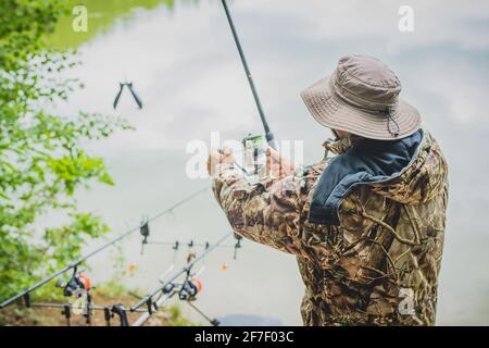 Pêche sportive pêcheur avec une canne et de la grenade alimentaire sur un lac. Vue arrière d'un pêcheur avec vêtements de camouflage et chapeau. Banque D'Images