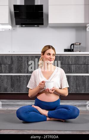 Femme de fitness heureuse enceinte Profitez d'une tasse d'eau ou de thé tout en étant assise sur le sol après un entraînement de yoga à la maison, femme en tenue sportive dans le salon, Banque D'Images