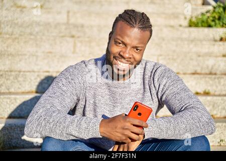 Gros plan d'un homme afro-américain assis dans les escaliers souriant, regardant la caméra, par une journée ensoleillée. Joyeux jeune Latino homme. Photo de haute qualité Banque D'Images