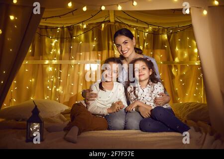 Portrait d'une mère et de deux filles assises dans une tente pour enfants douillette décorée de guirlandes. Banque D'Images