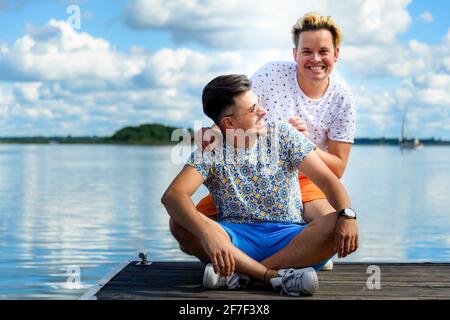 joyeux couple gay souriant assis sur une jetée en bois contre lac et ciel bleu Banque D'Images