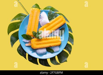 Délicieux popsicle de mangue fraîche servi avec des feuilles de menthe et des glaçons, un dessert frais fruité et sain, une glace de mangue isolée sur fond jaune Banque D'Images