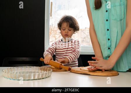 Petit enfant aidant la mère à rouler la pâte avec la broche à bord dans la cuisine à l'intérieur. Banque D'Images