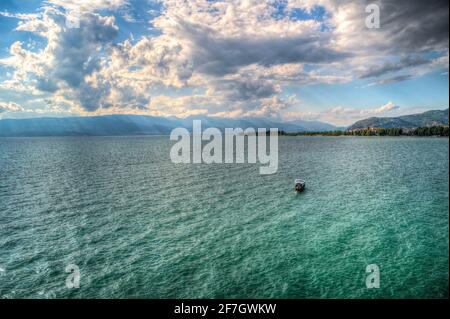 Bateau dans l'eau du lac, montagnes au loin, nuages épais dans le ciel. Bleu et vert avec beaucoup de détails. Banque D'Images