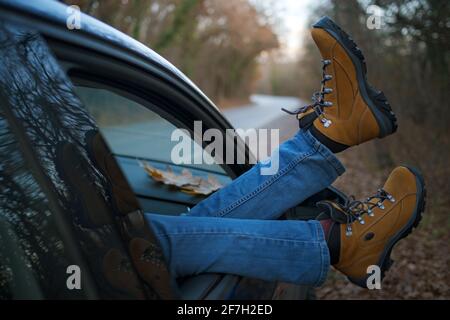 Femme pieds dans des bottes de randonnée jaune tendance sur la porte de voiture. Pieds à l'extérieur de la fenêtre au coucher du soleil forêt. Le concept de liberté de mouvement. Un week-end d'automne Banque D'Images