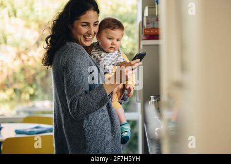 Femme envoyant un message texte sur son téléphone portable debout avec son bébé. Mère utilisant un téléphone portable tout en transportant son enfant à la maison. Banque D'Images