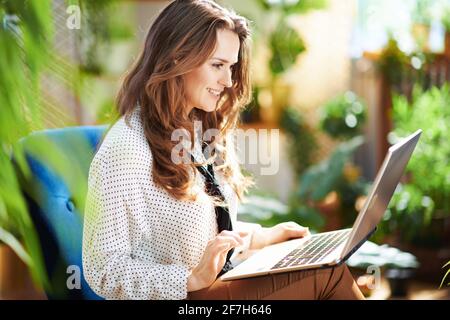 Green Home. Femme moderne heureuse avec de longs cheveux ondulés avec ordinateur portable dans la maison moderne en journée ensoleillée. Banque D'Images