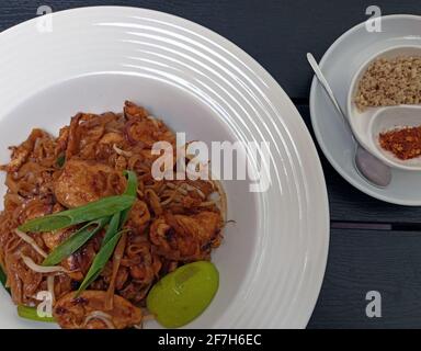 Papilez les nouilles thaïlandaises avec du poulet frit, des feuilles vertes mélangées, des cacahuètes hachées, des échalotes et un coin de citron vert sur une assiette blanche sur fond de bois servi Wi chaud Banque D'Images