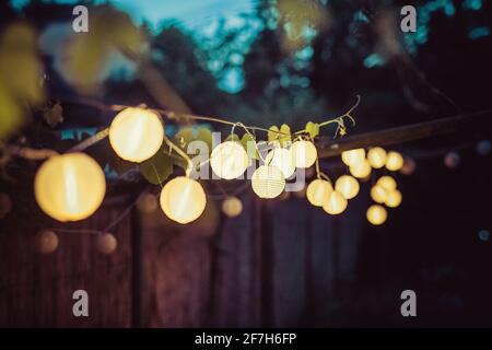 Rangée de lanternes rondes en papier dans un jardin sur un fond de chaume en bois la nuit. Décoration festive dans le jardin le soir. Banque D'Images