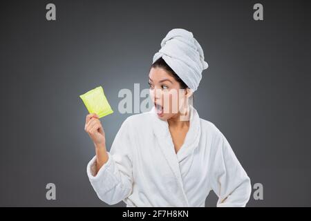 Ces jours du mois, les menstruations et les problèmes des femmes. Femme surprise dans un peignoir et une serviette enveloppée autour de la tête tenant une serviette sanitaire Banque D'Images