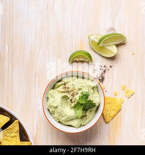 Trempette hummus à l'avocat et chips de maïs sur une table en bois blanc, vue sur le dessus, apéritif mexicain traditionnel dans un bol en céramique Banque D'Images