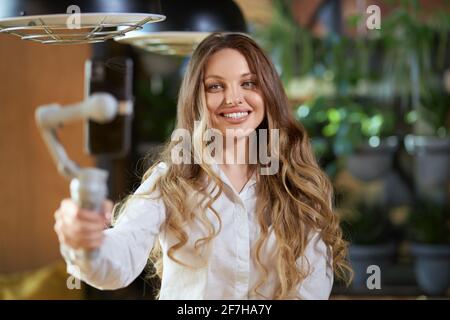 Portrait en gros plan d'une femme mignonne et souriante en chemise blanche tenant un bâton de selfie et communiquant avec les gens au téléphone. Concept de bon moment avec des abonnés en ligne parlant ou des amis. Banque D'Images