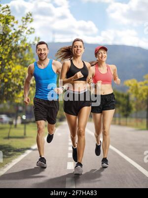 Portrait complet de deux jeunes femmes et d'un gars dans les vêtements de sport, courir sur une voie asphaltée Banque D'Images