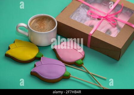 Pain d'épice sous forme de tulipes jaunes, roses et lilas avec une tasse de café blanc et une boîte de fête sur fond turquoise. Ambiance festive. Banque D'Images