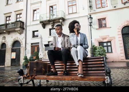 Beau homme en costume travaillant sur tablette numérique tandis que femme attrayante parlant sur mobile et écrivant sur presse-papiers. Deux collègues africains résolvant les problèmes de travail en dehors du bureau. Banque D'Images