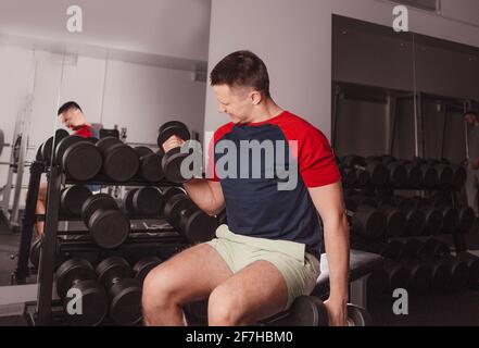 un homme soulève de lourdes haltères avec un visage tendu dans la salle de gym assis sur un banc. Banque D'Images