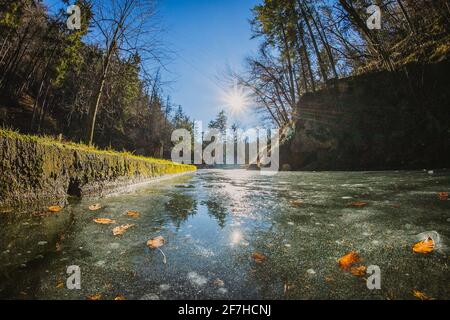 Détail de la surface de glace sur un petit lac ou un étang dans la nature entre les arbres d'hiver lors d'une journée ensoleillée avec des reflets de soleil et des stries visibles sur un ciel bleu clair. Banque D'Images