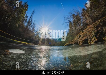 Détail de la surface de glace sur un petit lac ou un étang dans la nature entre les arbres d'hiver lors d'une journée ensoleillée avec des reflets de soleil et des stries visibles sur un ciel bleu clair. Banque D'Images