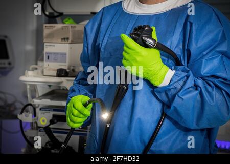 Médecin en vêtements de protection bleus avec des gants verts tient un endoscope en main. La lumière sur l'endoscope est allumée, le fond avec des utens de médecine Banque D'Images