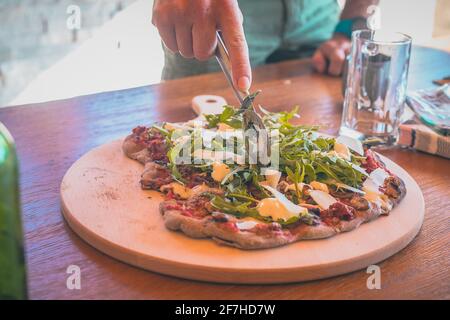 Délicieuse pizza maison sur une planche ronde en bois, fraîche d'une cheminée d'époque. Pizza en plein air. Homme en train de trancher des pizzas faites maison avec des ronds Banque D'Images