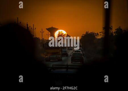 Trajet en soirée sur les routes africaines au Sénégal. Vue d'un bus vers le coucher du soleil et d'un château d'eau sur une route africaine animée. Banque D'Images