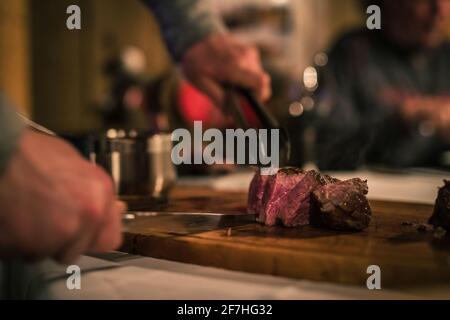 Un steak de filet moyen rare est coupé sur une planche de bois sur une table de restaurant. Des tranches de viande et des mains avec des couverts sont vues. Mise au point peu profonde. Banque D'Images