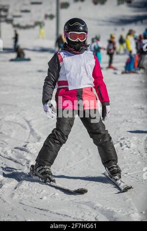 Enfant sur une piste de ski, portant un bavoir blanc de départ et un masque facial par temps ensoleillé. Enfant en cours de ski, debout sur des skis sans bâtons de ski. Banque D'Images