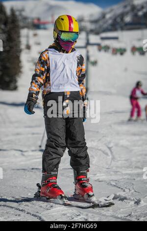 Enfant sur une piste de ski, portant un bavoir blanc de départ et un masque facial par temps ensoleillé. Enfant en cours de ski, debout sur des skis sans bâtons de ski. Banque D'Images