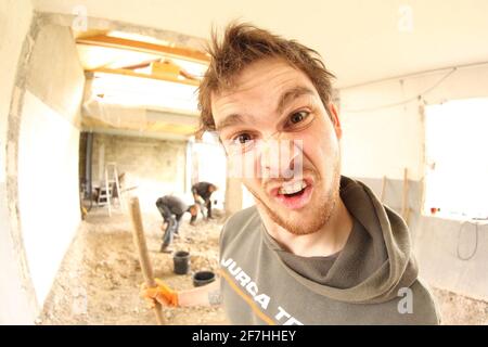 Un travailleur qui fait un visage stupide tout en rénovant une maison. Autres travailleurs en arrière-plan. Vue fisheye. Banque D'Images